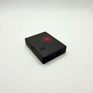 May include: A rectangular black plastic case with a textured finish. The top surface features a red circular target graphic and three small vertical ventilation slits. A small port opening is visible on the side of the compact electronic device.