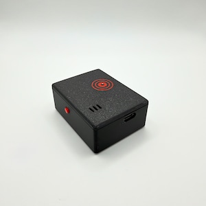 Könnte beinhalten: Ein rechteckiges, schwarzes elektronisches Gerät mit strukturierter Oberfläche. Die Oberseite zeigt ein rotes Zielscheiben-Motiv und ein kleines Lautsprechergitter. Ein roter Knopf befindet sich an der Seite, ein USB-C-Ladeanschluss ist vorne.