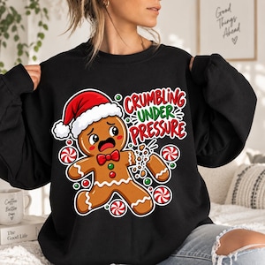 May include: A black crewneck sweatshirt featuring a graphic of a distressed gingerbread person wearing a Santa hat, with one arm breaking off. The text reads "CRUMBLING UNDER PRESSURE" in red and green, surrounded by red and white peppermint candies.
