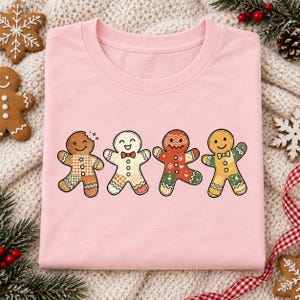 Può includere: Una t-shirt rosa chiaro a girocollo con una stampa grafica di quattro biscotti di pan di zenzero in fila. Ogni biscotto presenta un motivo patchwork unico nei toni del marrone, rosso, verde e bianco, con dettagli decorativi di glassa.