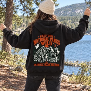 May include: A black hooded sweatshirt featuring a graphic on the back with the text KEEP OUR NATIONAL PARKS WILD OR THEY'LL FOLLOW YOU HOME. The design includes illustrations of Bigfoot, an alien, a sea serpent, and ghosts in a forest.