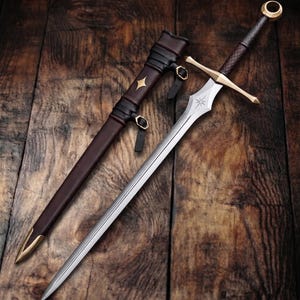 May include: A silver-toned steel sword with a gold-colored crossguard and pommel, featuring a star-shaped engraving on the blade. The sword is accompanied by a dark brown leather scabbard with gold-colored metal accents and black leather straps.