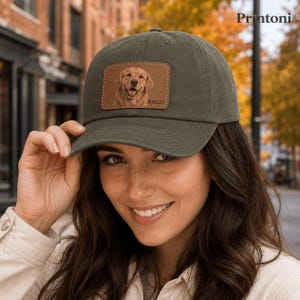 May include: An olive green baseball cap featuring a rectangular brown leather patch on the front. The patch displays an engraved illustration of a golden retriever dog with the name BAILEY printed in capital letters next to the dog's image.
