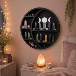May include: A circular black wooden wall shelf shaped like a crescent moon, featuring multiple horizontal tiers holding various polished crystal points and spheres. A white triple moon symbol is printed on the center back panel of the shelf.