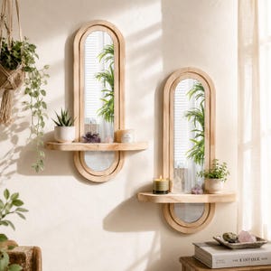 May include: Two arched wall mirrors with light wood frames, each featuring an integrated horizontal shelf. The mirrors reflect indoor plants and light, while the shelves hold small potted plants, candles, and crystals against a plain wall.