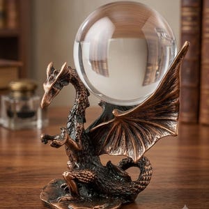 Puede incluir: Una figura de dragón color bronce con escamas texturizadas y grandes alas extendidas. El dragón está posado sobre una base y sostiene una gran esfera de cristal transparente sobre su espalda, creando una pieza decorativa con acabado metálico.