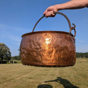 May include: A large, round, hammered copper pot with a polished metallic finish. The vessel features a dark, textured iron swing handle attached to the sides with decorative metal rivets and a curved hook detail on one side of the rim.