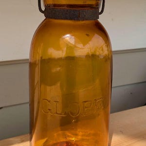 Puede incluir: Un frasco de vidrio de color ámbar con la palabra GLOBE grabada en relieve en el frente. El frasco cuenta con una tapa de metal oscuro y oxidado con un asa de alambre sujeta a una banda metálica que rodea el cuello del recipiente.
