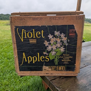 May include: A rectangular wooden crate featuring a black label with yellow text reading Violet Brand Apples. The label displays a bouquet of purple violets and text including Perham Fruit Corporation, Yakima, Washington, and Contents One U.S. Bushel.