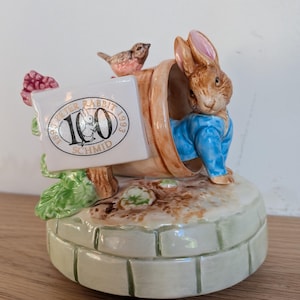 May include: A ceramic music box featuring a brown rabbit in a blue jacket emerging from a terracotta pot. A white sign attached to the pot reads 1893 Peter Rabbit 1993 100 Schmid. A small bird sits on the pot, which rests on a green brick base.