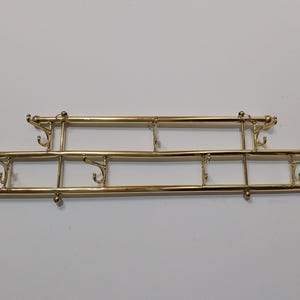 May include: A polished gold-tone metal wall-mounted coat rack featuring two horizontal bars connected by vertical supports, with multiple curved hooks extending from the bars for hanging items in an organized arrangement.