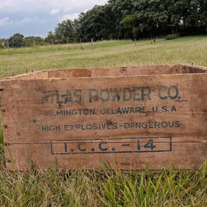 May include: A rectangular wooden crate with weathered brown panels and visible metal nails. The front features black stenciled text reading ATLAS POWDER CO. WILMINGTON, DELAWARE, U.S.A., HIGH EXPLOSIVES - DANGEROUS, and I.C.C.-14, with an open top design.
