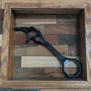 May include: A black metal multi-tool featuring a bottle opener head with a curved hook and a hexagonal cutout, connected to a long handle that terminates in a large hexagonal opening, designed for various mechanical or utility tasks.