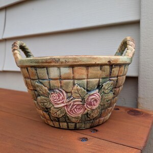 May include: A round ceramic basket with two handles, featuring a textured woven pattern in shades of tan, brown, and green. The exterior is decorated with a central relief design of three pink roses surrounded by green leaves and stems.
