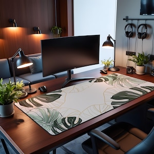 May include: A rectangular desk mat featuring a white background with a repeating pattern of large, dark green monstera leaves outlined in thin gold lines. The mat is positioned on a dark wood desk surface beneath a curved computer monitor.