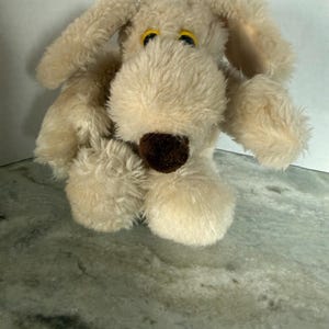 May include: A plush dog toy with cream-colored shaggy fur, long floppy ears, and a dark brown nose. The stuffed animal features bright yellow eyes with black pupils, a rounded snout, and a soft, textured appearance throughout its body.