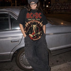 Puede incluir: Una camiseta negra de manga corta con un gráfico rojo y amarillo de AC/DC PWR UP TOUR que incluye la ilustración de un guitarrista, combinada con una gorra negra, pantalones cargo negros y zapatillas deportivas en blanco y negro.