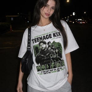 Może przedstawiać: Biały t-shirt z krótkim rękawem z zielono-czarnym nadrukiem graficznym z tekstem "TEENAGE KIX", "SEASON FIVE EPISODE TWO OF 'THE BOYS'", "WRITTEN BY GARTH ENNIS" oraz "IF YOU CAN'T BEAT 'EM, JOIN 'EM. THAT'S THE FUCKING VIBE."
