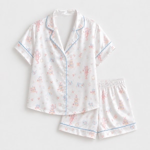 May include: A white short-sleeve pajama set featuring a button-down shirt with a notched collar and matching shorts. The fabric is decorated with a repeating pattern of small pink and blue illustrations of cats and cakes, accented by light blue piping.