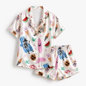 May include: A two-piece pajama set in cream featuring a repeating print of skeletons on beach chairs, pink surfboards, jack-o'-lanterns, ice cream cones, watermelon slices, and umbrellas. The top has a collared button-down front and short sleeves.