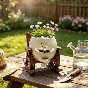 May include: A cream-colored ceramic planter shaped like a character sitting in a brown wooden rocking chair, holding an open book. The planter contains green grass and small white and pink daisy-like flowers, with a smooth, rounded face design.