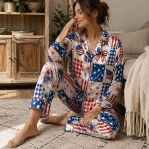 Könnte beinhalten: Ein zweiteiliger Schlafanzug mit Patchwork-Muster in Rot, Weiß und Blau. Das Design zeigt amerikanische Flaggen, Sterne, Vichy-Karos, Feuerwerk und Schleifen auf hellem Grund. Das Oberteil hat einen Kragen und eine durchgehende Knopfleiste.