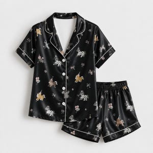 May include: A black pajama set consisting of a short-sleeved button-down shirt with a notched collar and matching shorts. The fabric features a repeating pattern of small unicorns wearing party hats, flowers, and bows with white piping trim.