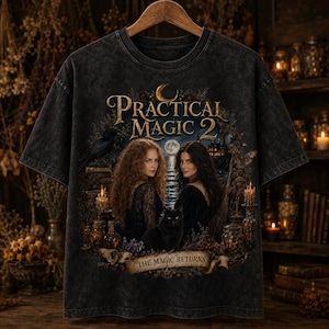 以下が含まれることがあります： チャコールグレーのミネラルウォッシュ加工Tシャツ。2人の女性、黒猫、カラスのグラフィックプリントが施されています。テキストには「PRACTICAL MAGIC 2」と「THE MAGIC RETURNS」と記され、キャンドル、月、家が描かれたダークで雰囲気のあるデザインです。