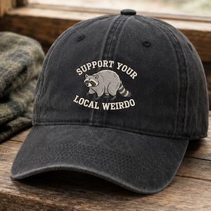 May include: A charcoal black baseball cap featuring an embroidered graphic of a raccoon in the center. The text "SUPPORT YOUR LOCAL WEIRDO" is arched around the raccoon illustration in a light cream color. The cap has a curved brim and eyelets.