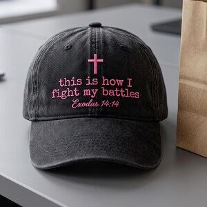 May include: A charcoal black baseball cap featuring embroidered pink text that reads "this is how I fight my battles" with "Exodus 14:14" underneath and a small pink cross symbol centered above the lettering on the front panel.