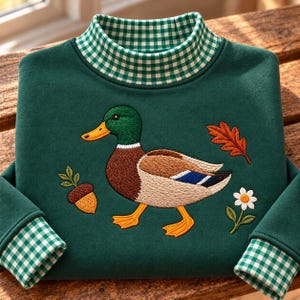 May include: A dark green long-sleeve sweatshirt featuring a mock neck collar with a white and green gingham pattern. The center displays an embroidered mallard duck, an acorn, a single orange leaf, and a white daisy with a yellow center.