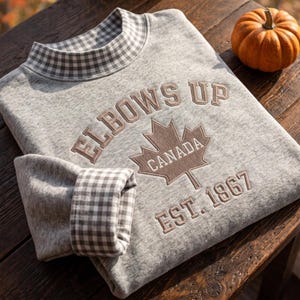 May include: A heather gray sweatshirt featuring a mock neck collar with a gray and white gingham pattern. The chest displays embroidered text reading ELBOWS UP, a maple leaf with CANADA, and EST. 1867. The sleeve cuffs also feature the gingham pattern.