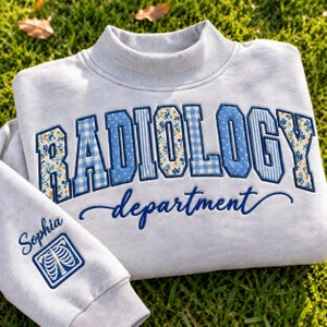 May include: A light gray mock neck sweatshirt featuring the word RADIOLOGY in large, blue-bordered letters with varied floral, gingham, and polka dot patterns. Below, the word department is embroidered in blue script, with Sophia and a rib cage icon.