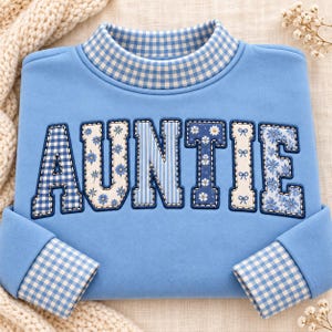 May include: A light blue sweatshirt featuring a mock neck collar and cuffs in a blue and white gingham pattern. The word AUNTIE is appliqued across the chest in large letters, each filled with varying blue and white floral, striped, and gingham fabrics.
