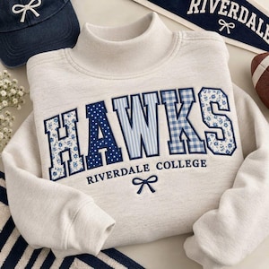 May include: A heathered light gray mock neck sweatshirt featuring the word HAWKS in large, blue-toned patterned letters. Below the text, the words RIVERDALE COLLEGE are embroidered in blue, accompanied by a small blue bow icon centered underneath.