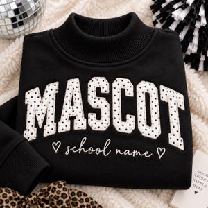 May include: A black sweatshirt with a ribbed mock neck collar. The word MASCOT is appliqued in white fabric with black polka dots. Below the text, the words school name are embroidered in white cursive, flanked by two small white heart outlines.