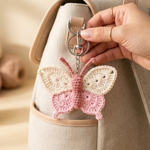 May include: A crochet butterfly keychain featuring a dusty rose body and antennae, with upper wings in cream and lower wings in dusty rose. The butterfly is attached to a silver-toned metal lobster clasp and split ring, hanging from a beige backpack.