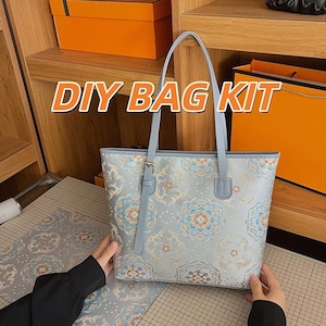 Puede incluir: Un bolso tote de color azul claro con un patrón floral repetitivo estilo mandala en tonos naranja, blanco y azul oscuro. El bolso tiene dos correas delgadas para el hombro y un detalle de correa vertical decorativa en el frente. El texto DIY BAG KIT es visible.