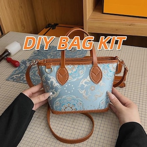 Op de afbeelding: Een DIY BAG KIT met een lichtblauwe handtas met lichtbruine lederen afwerking en handvatten. De tas heeft een herhalend patroon van gestileerde herten en bloemmotieven. Het bevat een bijpassende lange schouderriem en een ritssluiting aan de bovenkant.