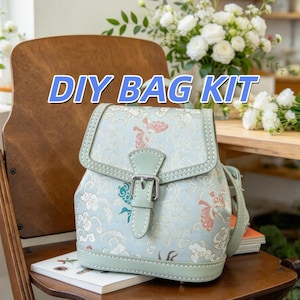 May include: A light green backpack featuring a patterned design of butterflies and clouds in shades of pink, white, and blue. The bag has a front flap with a silver metal buckle closure and stitched edges. The text DIY BAG KIT is visible above.