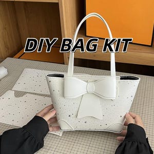 Puede incluir: Un bolso de mano DIY BAG KIT blanco con un patrón de estrellas y un gran lazo central. El bolso tiene dos asas superiores y esquinas reforzadas. El kit incluye piezas de material blanco precortadas con un estampado de estrellas a juego.