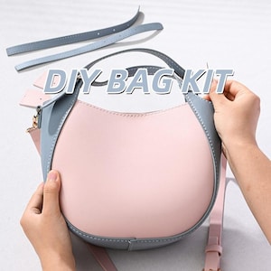 Puede incluir: Un kit de bolso DIY que incluye un bolso de mano semicircular con cuerpo rosa claro y ribete azul pizarra. El kit contiene correas a juego en azul pizarra y una correa larga rosa para el hombro. El texto DIY BAG KIT aparece en blanco.