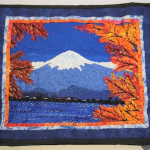May include: A blue and white embroidered wall hanging featuring a mountain range with a lake in the foreground. The mountain range is surrounded by orange and red leaves.