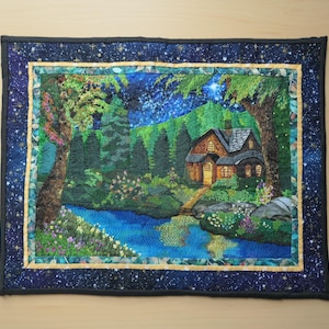 May include: A quilted wall hanging depicting a serene landscape with a house nestled among trees and a river. The night sky is filled with stars. The border is dark blue with a starry pattern, and the inner border is gold.
