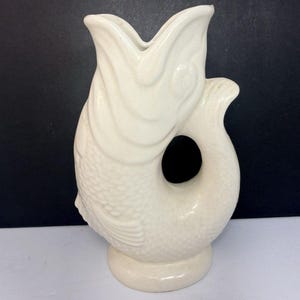 May include: A cream-colored ceramic pitcher shaped like a fish, featuring a wide open mouth at the top, a circular handle formed by the curved tail, and detailed scale textures covering the body, standing on a circular base.