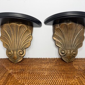 May include: A pair of wall-mounted shelves featuring semi-circular black tops and ornate, gold-toned brackets shaped like stylized scallop shells with scrolled bases. The design incorporates detailed fluting and symmetrical decorative elements.