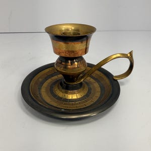 Puede incluir: Un candelabro de latón con una base circular que presenta anillos concéntricos oscuros y dorados. El tallo central sostiene una copa ancha y un asa curva se extiende desde el lateral. La superficie metálica muestra una mezcla de tonos pulidos y oscuros.