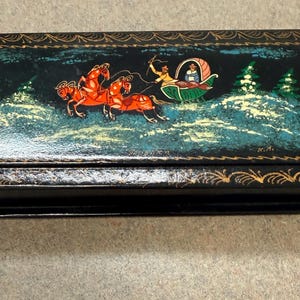 May include: A rectangular black lacquered box featuring a hand-painted scene on the lid of a troika with three red horses pulling a carriage through a snowy landscape with green trees. The edges are decorated with a gold-coloured leaf pattern.