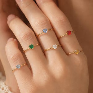 May include: A collection of six thin gold rings, each featuring a small heart-shaped gemstone in a different color, including light blue, red, green, clear, yellow, and pale blue, worn across multiple fingers on a single hand.