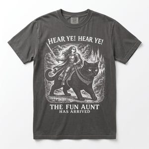 Puede incluir: Una camiseta gris carbón con un estampado gráfico blanco de una persona con armadura montada sobre un gato negro grande. El texto dice HEAR YE! HEAR YE! en la parte superior y THE FUN AUNT HAS ARRIVED en la parte inferior, con un castillo.